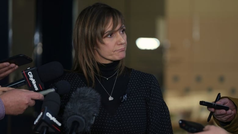 a woman stands and looks to her left. there are at least 7 microphones or recording devices pointed at her.