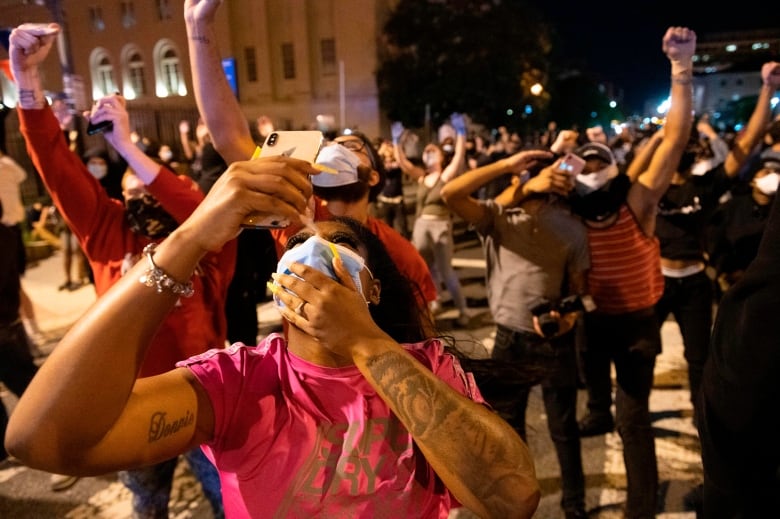 Several people are shown standing outside on a street in a nighttime photo, looking up at the sky. They are wearing COVID-19 masks and some are holding phones up.