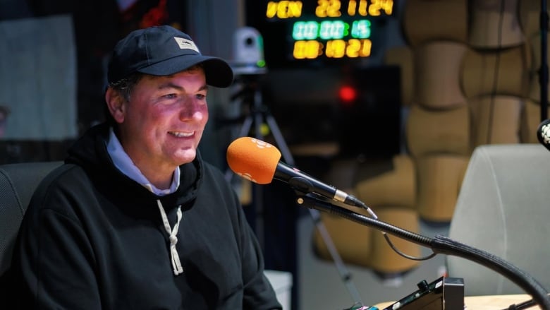 A man in a baseball hat wearing a sweater smiling while sitting in front of an orange microphone. 