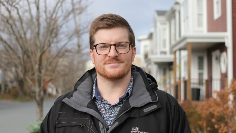 A man with glasses, a beard and mustache wears a coat and stands on a sidewalk next to a row of houses. He has a small smile.