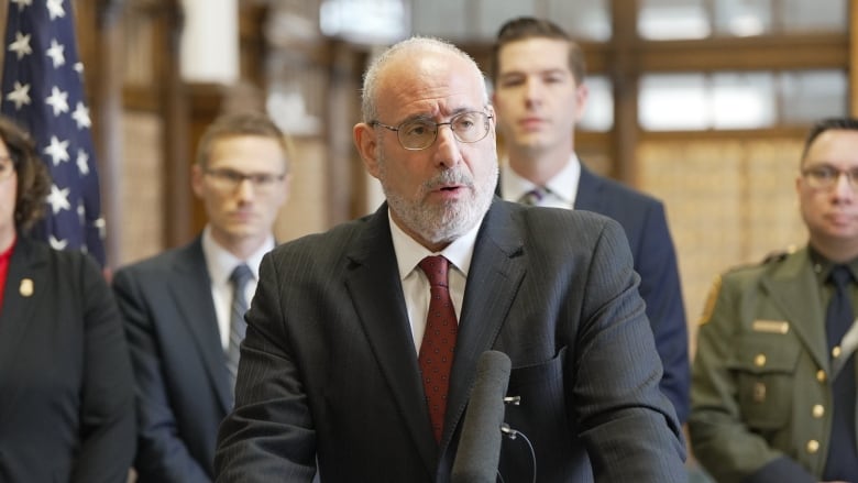 A man in a suit speaks in front of a podium with others, including two police officers, standing behind him.