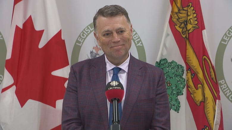 A man wearing a purple suit and blue tie stands in front of the Canadian and Prince Edward Island flags.