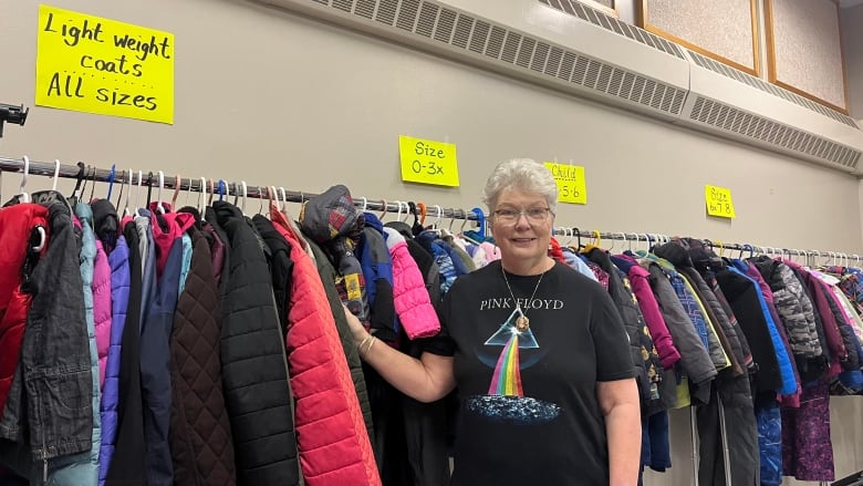 Woman with glasses stands beside rack of coats. 