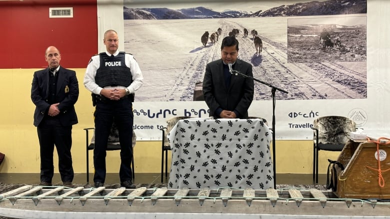 Man in black suit speaks into a microphone. A dog sled and qulliq are viisible