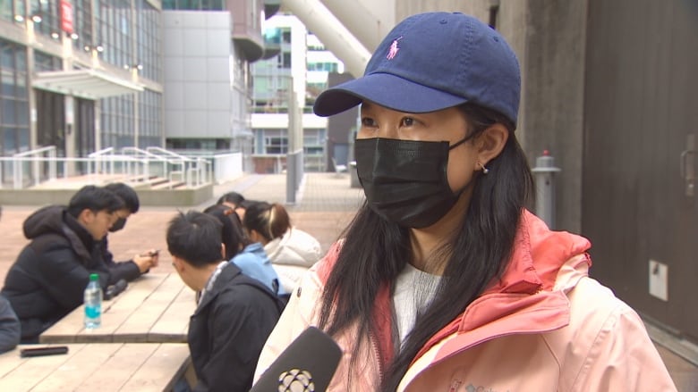 A woman in a cap and wearing mask can be seen speaking into a mic.