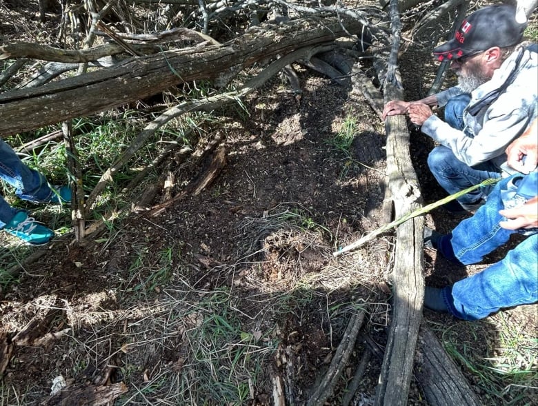 A man crouches down, looking at the ground in a treed area. 