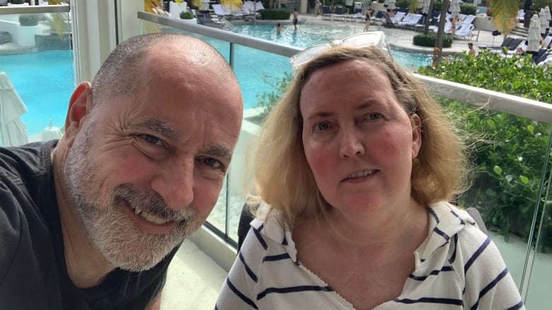 A balding man with a grey goatee smiles for the camera while a woman in a white-striped top looks quizzically into the lens.