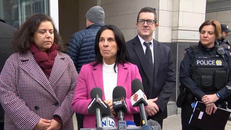 A white woman with black hair in a pink blazer speaks into several microphones.