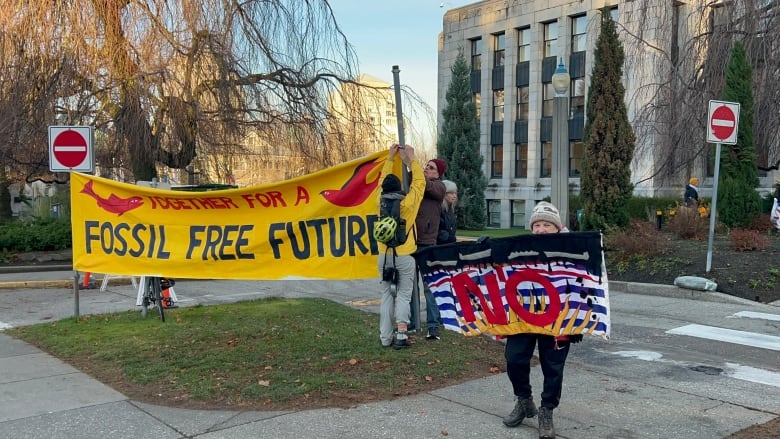 People put up a yellow banner that says 'Together for a fossil free future' while a woman holds up a B.C. provincial flag with 'NO' written on it.