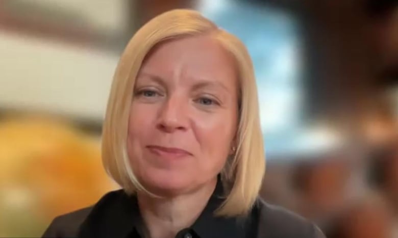  A woman with chin length blond hair, wearing a black collared shirt, is smiling and looking into the camera