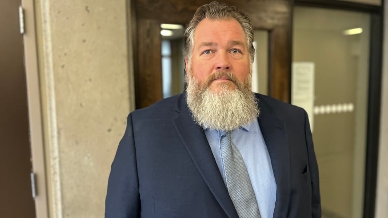 A man with a beard in a suit and tie stands in a hallway.